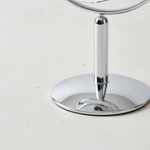 Espelho de aumento para maquiagem, redondo, detalhe da base de mesa cromada com reflexo em superfície clara.