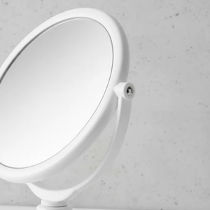 Espelho de aumento para maquiagem, redondo, de mesa, acabamento branco, em ambiente de banheiro, com foco na lateral do espelho.