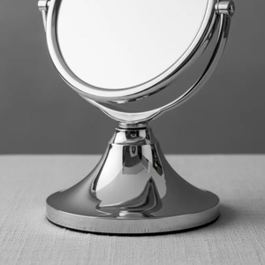 Espelho de aumento para maquiagem, redondo, de mesa, acabamento cromado, sobre superfície neutra, destaque para base metálica.