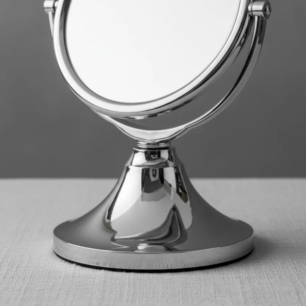 Espelho de aumento para maquiagem, redondo, de mesa, acabamento cromado, sobre superfície neutra, destaque para base metálica.