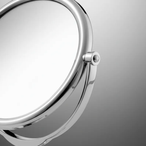 Espelho de aumento para maquiagem, redondo, acabamento cromado, em ambiente neutro, destaque para o detalhe da moldura metálica.